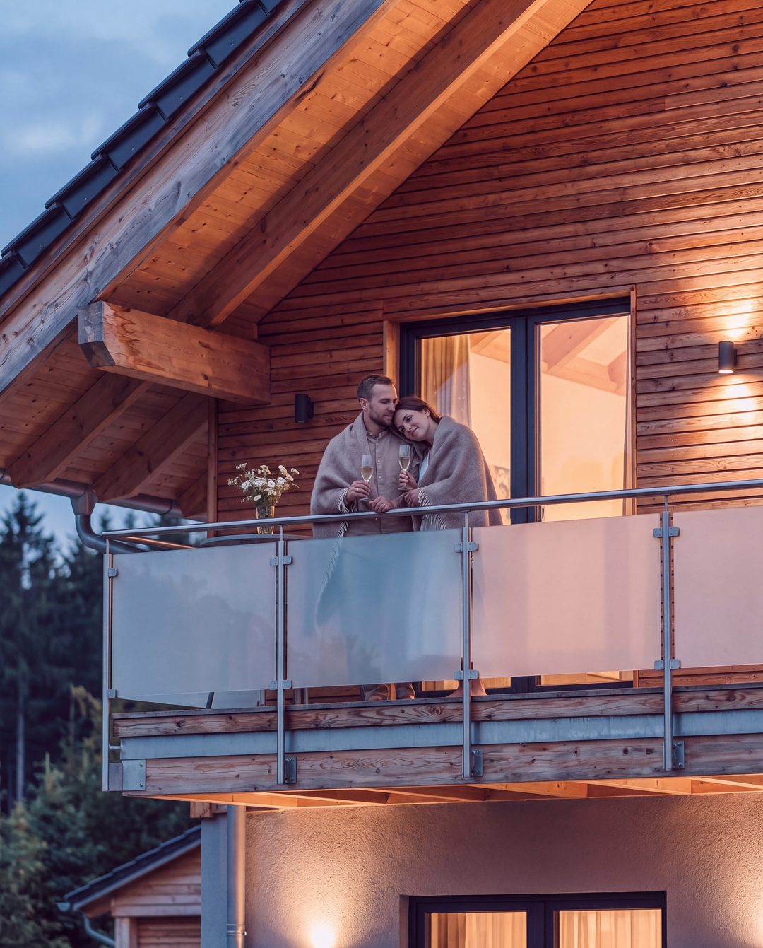 Una pareja envuelta en una manta de lana con copas de champán en el balcón de un chalet a la hora azul, imagen ilustrativa