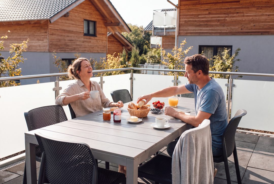 Una pareja desayuna riendo en la terraza de un chalet, con panecillos, fresas y zumo de naranja sobre la mesa; imagen ilustrativa