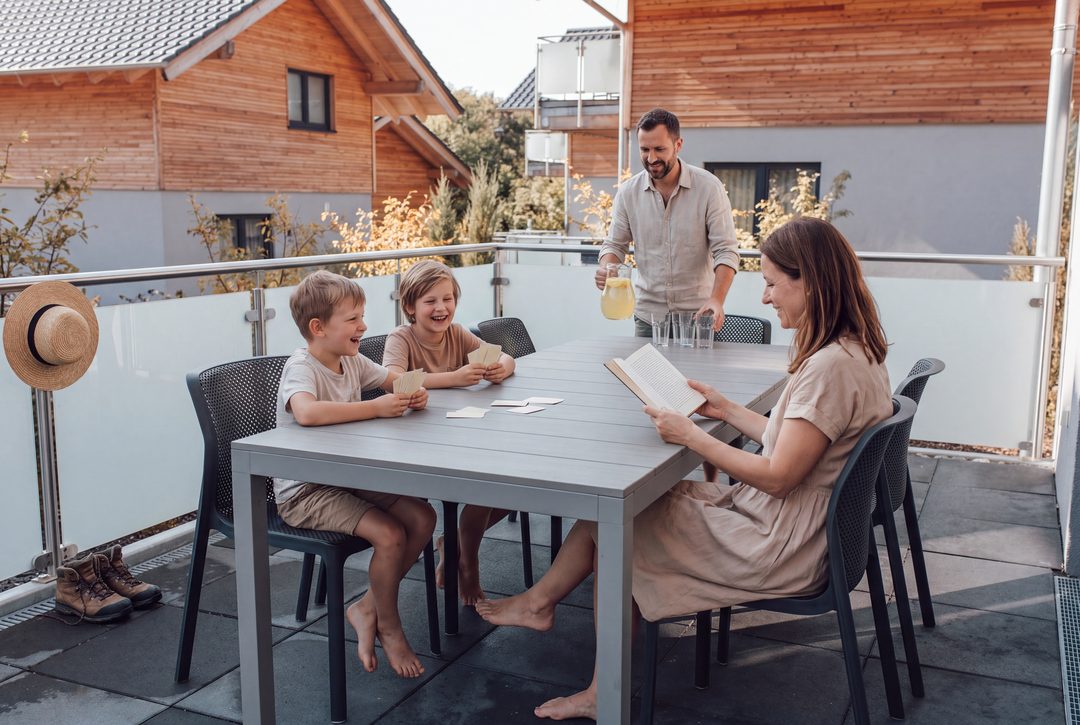 Una familia en la terraza de un chalet: dos niños juegan a las cartas riendo, la madre lee y el padre les trae limonada; imagen ilustrativa