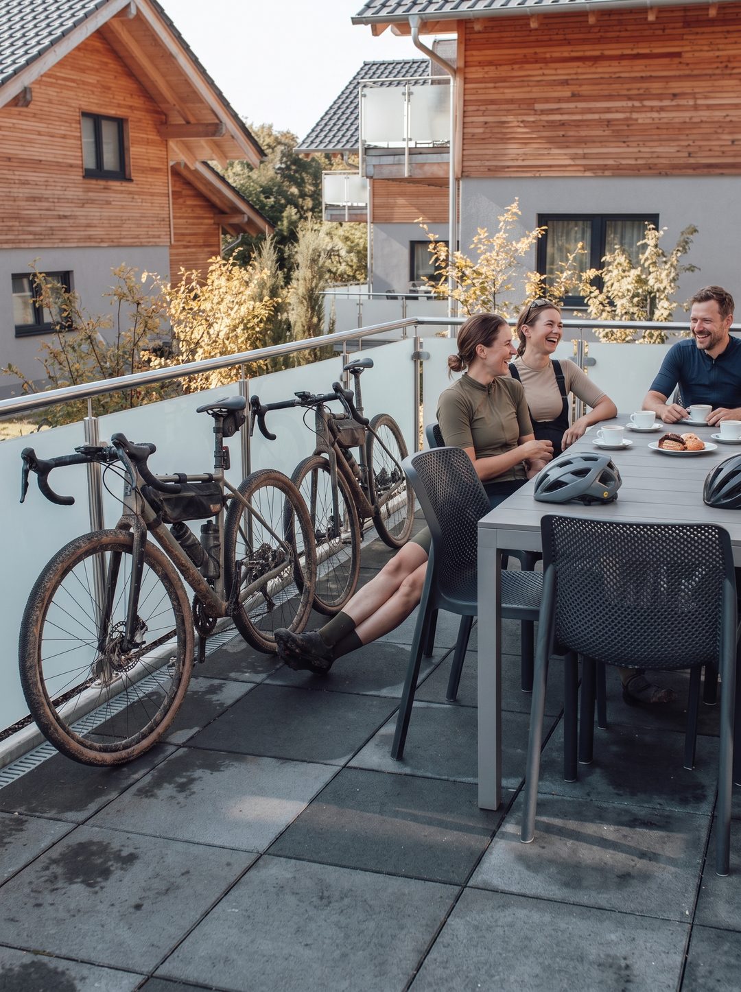 Hochformat: Gravelbiker lachen beim Kaffee auf der Chalet-Terrasse, zwei staubige Gravelbikes lehnen am Geländer, Symbolbild