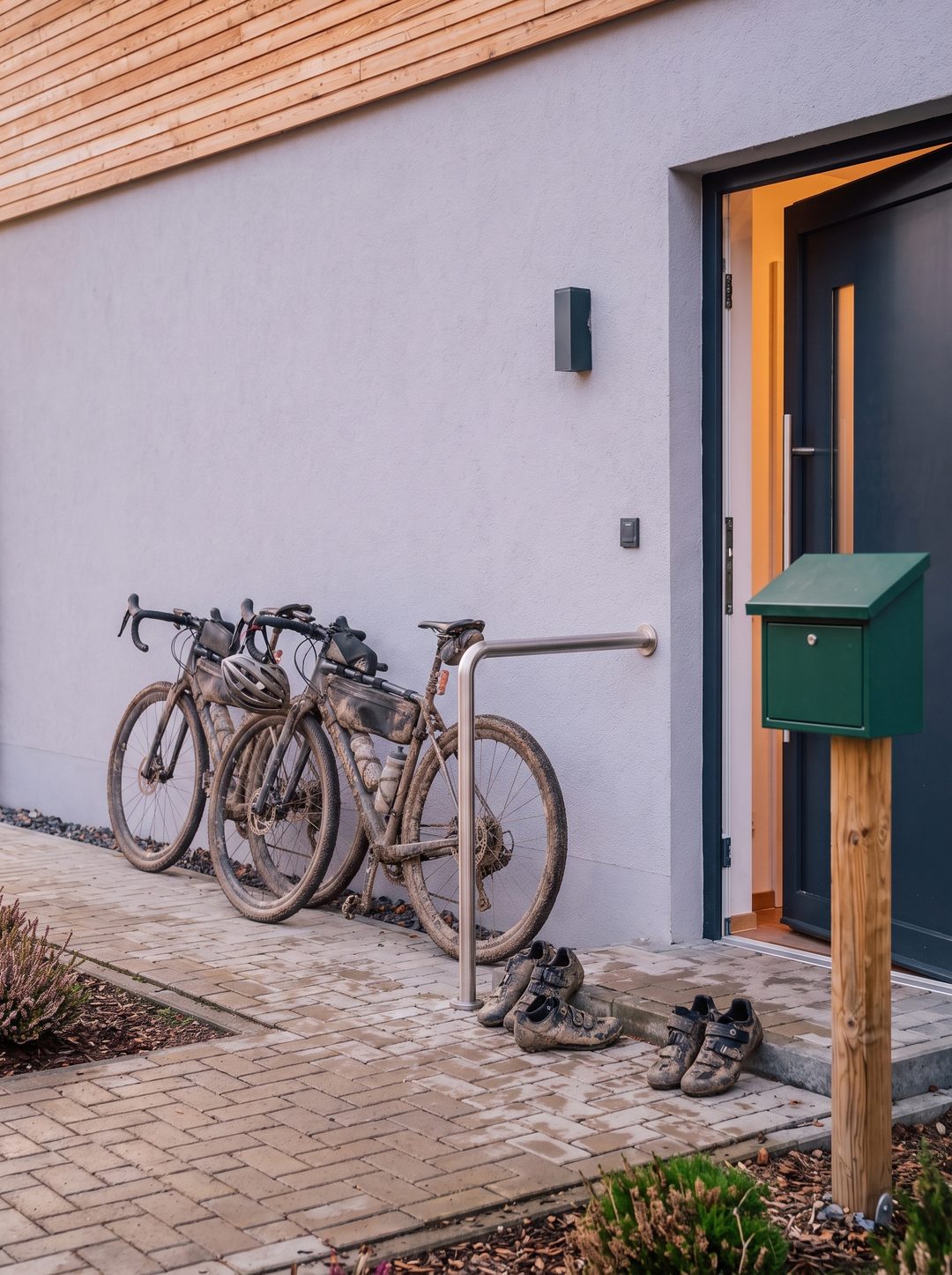 Hochformat: zwei verschlammte Gravelbikes an der Hauswand neben Haustür und Briefkasten eines Chalets, Symbolbild