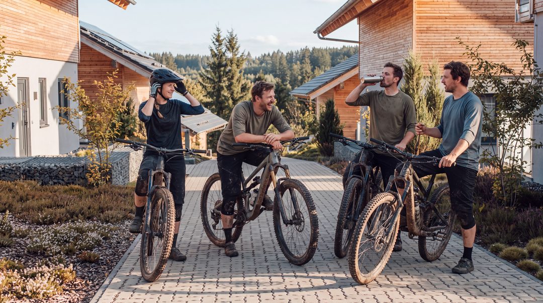 Fire mountainbikere med snavsede cykler og knæbeskyttere griner efter turen på stien mellem træchaletterne, symbolbillede