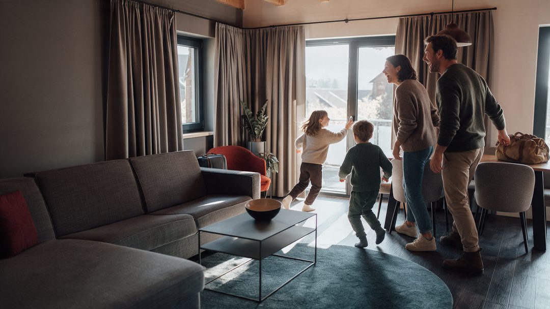 En familie ankommer til stuen i et chalet: Børnene løber hen til terrassedøren, forældrene griner, symbolbillede