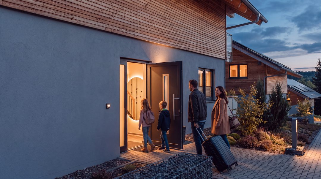 Una familia con maletas con ruedas entra en un chalet al atardecer por la puerta principal, que está abierta y tiene una cálida iluminación; imagen ilustrativa