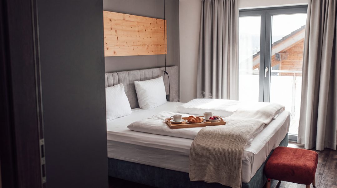 Frisch bezogenes Boxspring-Doppelbett mit weißer Bettwäsche, Strickdecke und Frühstückstablett mit zwei Tassen Kaffee im Morgenlicht eines Chalet-Schlafzimmers, Symbolbild