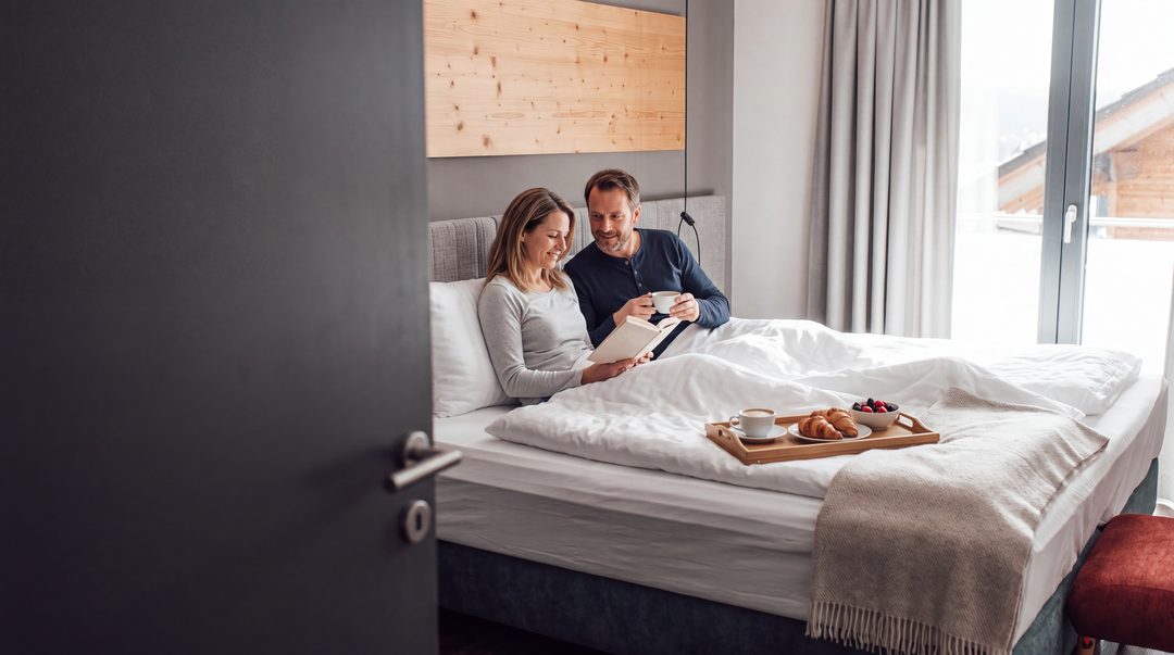 Una pareja está sentada por la mañana en la cama doble con somier recién hecha de un dormitorio del chalet; ella lee, él tiene una taza de café y una bandeja de desayuno sobre la colcha; imagen ilustrativa