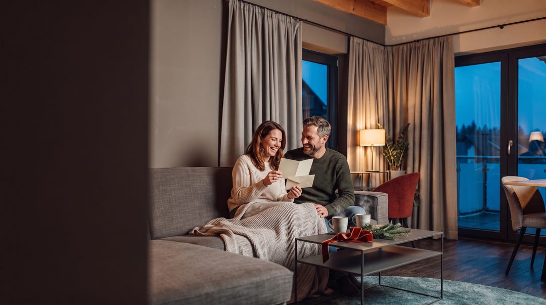 Una pareja lee juntos en el sofá de un chalet una tarjeta que viene en un sobre de regalo; imagen ilustrativa