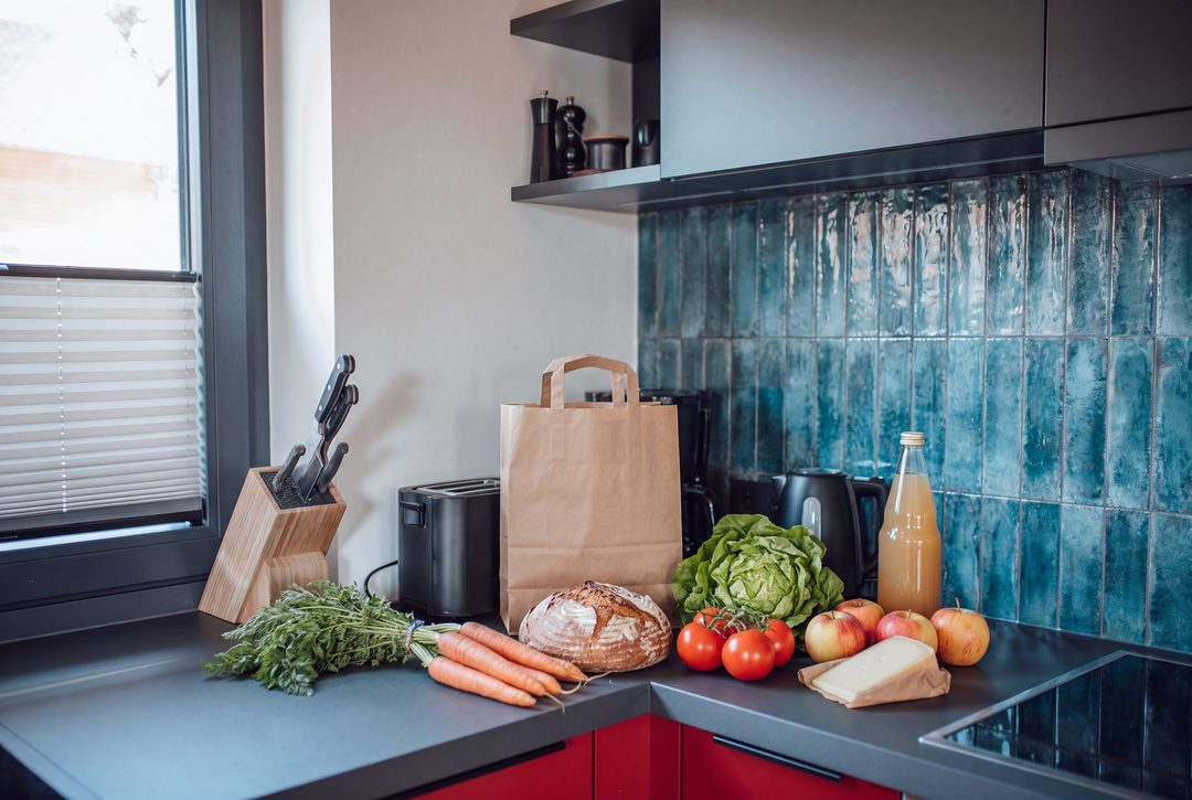 Courses déballées sur le plan de travail de la cuisine d'un chalet : sac en papier, pain, carottes, salade, tomates, pommes, fromage de montagne et jus de pomme devant un carrelage bleu, image d’illustration
