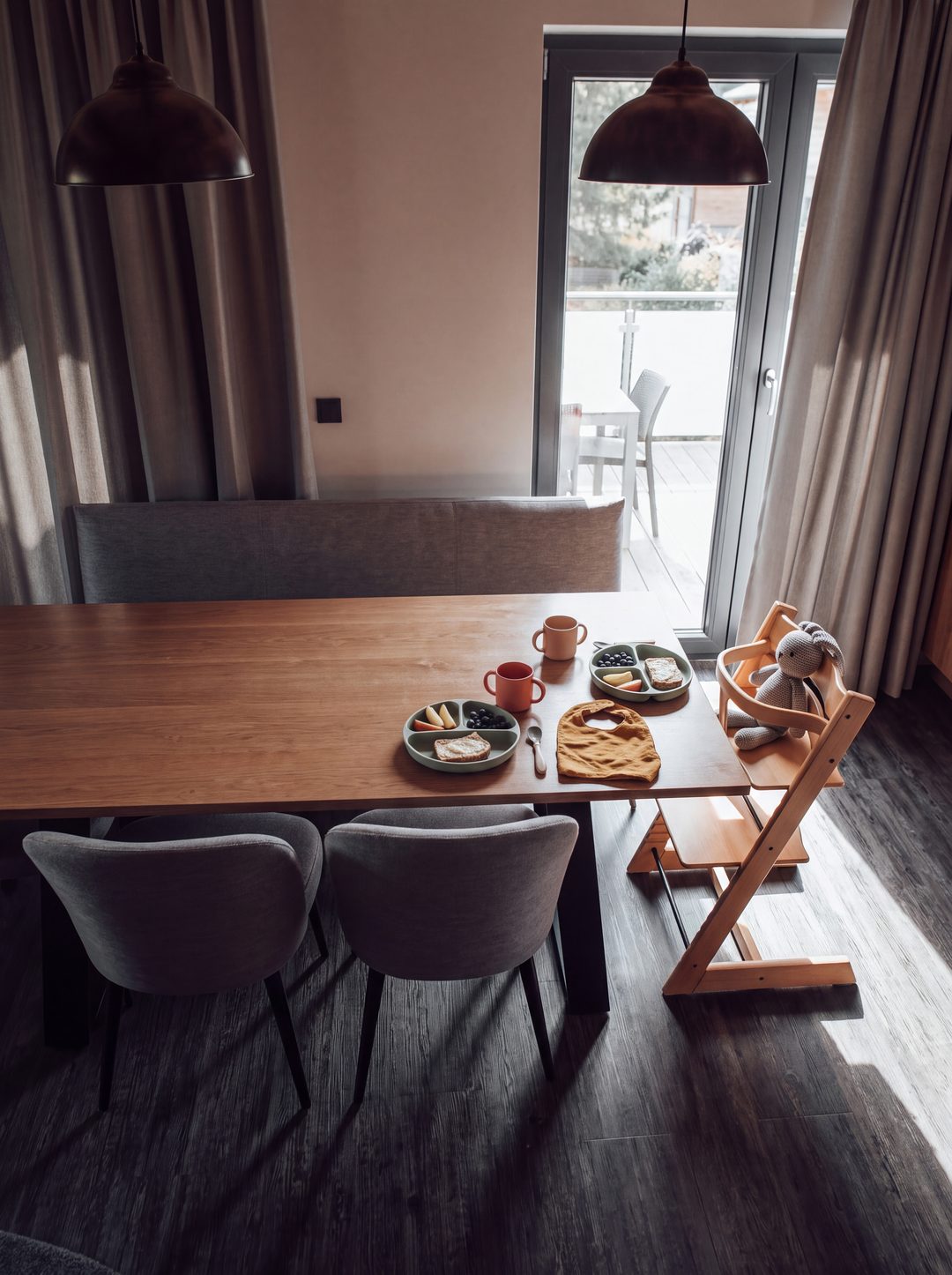 Træbørnestol med strikket kanin ved spisebordet i et chalet, foran den to børnetallerkener med frugt og brød, to krus og et hagesmæk, morgenlys gennem altandøren, symbolbillede
