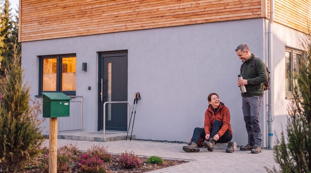 Un couple de randonneurs de retour au chalet à la lumière du soir : elle est assise devant la porte d'entrée et délace ses chaussures de randonnée, tandis qu'il tient le thermos en riant ; image d’illustration