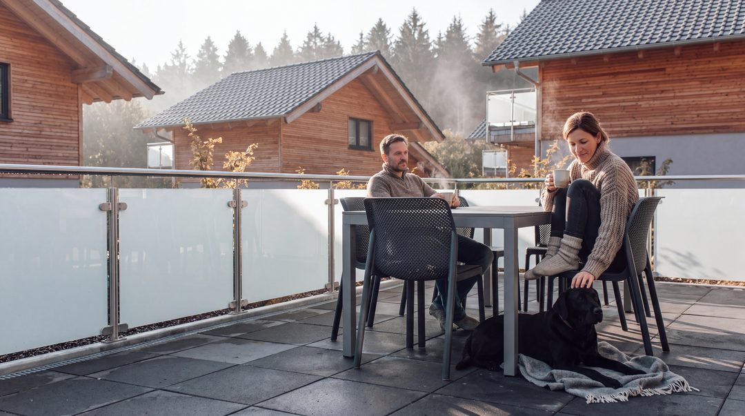 Una pareja toma café a primera hora de la mañana en la terraza de un chalet; el labrador negro está tumbado en su manta junto a ellos, imagen ilustrativa