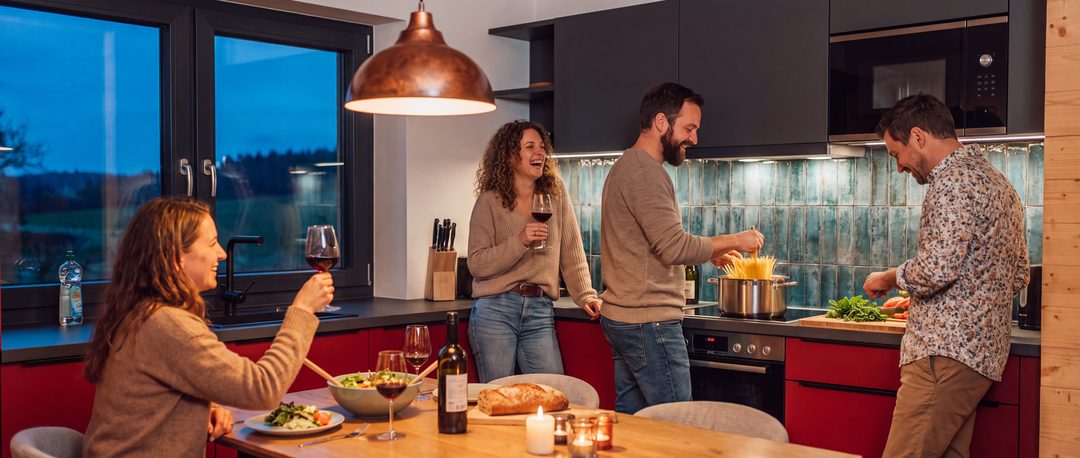 Cuatro amigos cocinan pasta por la noche, riendo, en la cocina de un chalet con fachadas rojas; una amiga levanta su vaso en la mesa del comedor, imagen ilustrativa