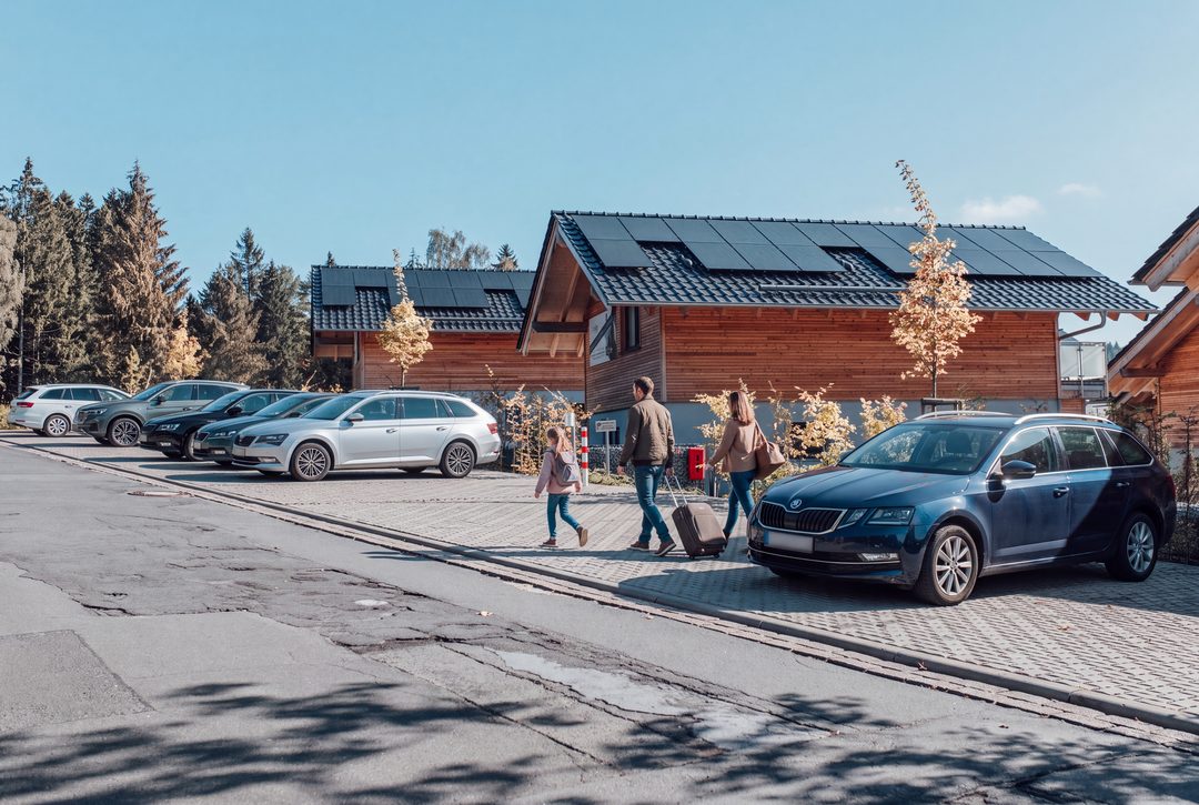 Aparcamiento delante de los chalets revestidos de madera con coches aparcados; una familia se dirige con maletas hacia el camino que lleva a los chalets, imagen ilustrativa