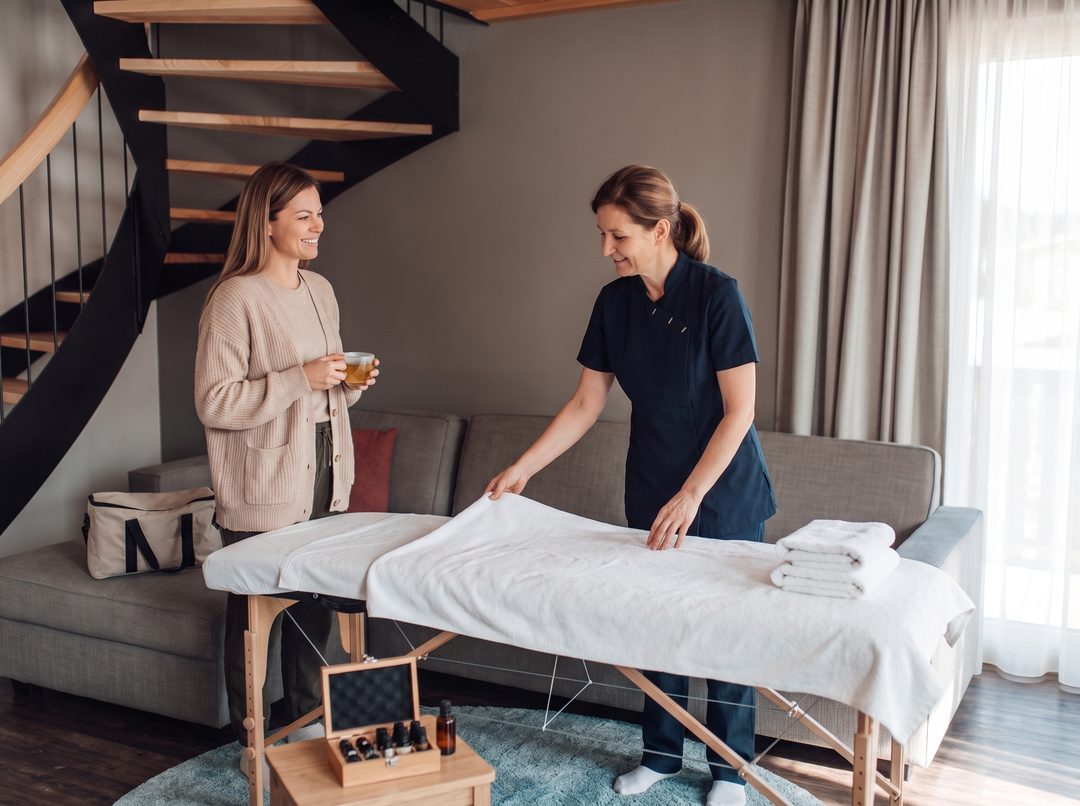 En terapeut sætter massagelejet op i stuen i et chalet, mens en gæst med en tekop i hånden snakker med hende – symbolbillede