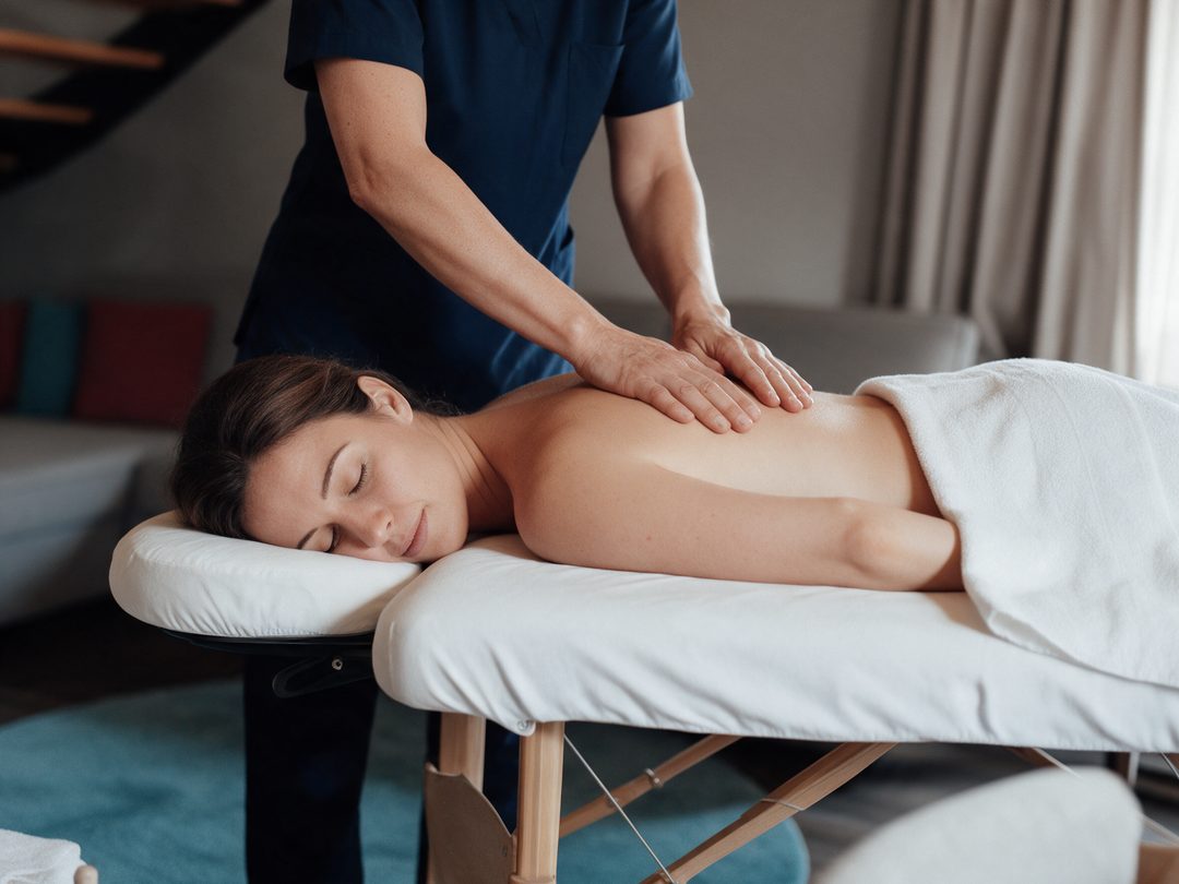 Las manos de la terapeuta durante un masaje clásico de espalda; la huésped está tumbada relajada en la camilla con los ojos cerrados, imagen ilustrativa