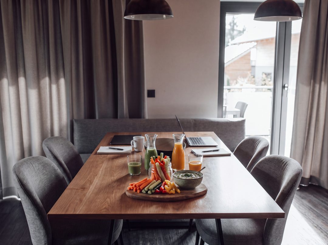 Pause under et møde ved spisebordet i et chalet: Grøntsagssticks med dip, grønne og orange smoothies, samt notesblokke og en bærbar computer, symbolbillede