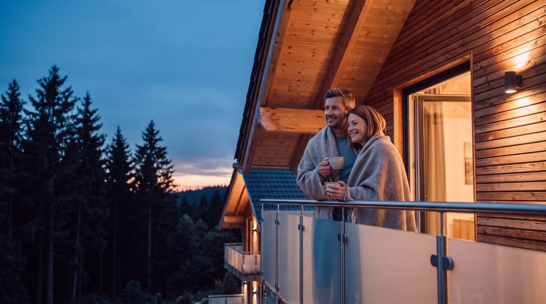 Paar in Decken gehüllt mit Tee auf dem Balkon eines Holzchalets zur Blauen Stunde, Tannen im Hintergrund, Symbolbild