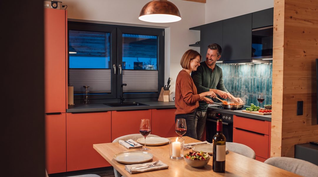 Una pareja cocina por la noche, riendo, en la cocina de un chalet con frentes rojos; dos copas de vino tinto, la mesa puesta con una vela, imagen ilustrativa