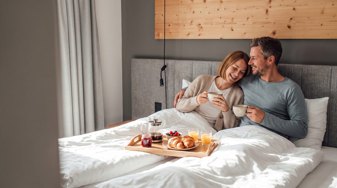 Una pareja desayuna café y cruasanes en la cama box spring de un dormitorio del chalet, a la luz de la mañana; imagen ilustrativa