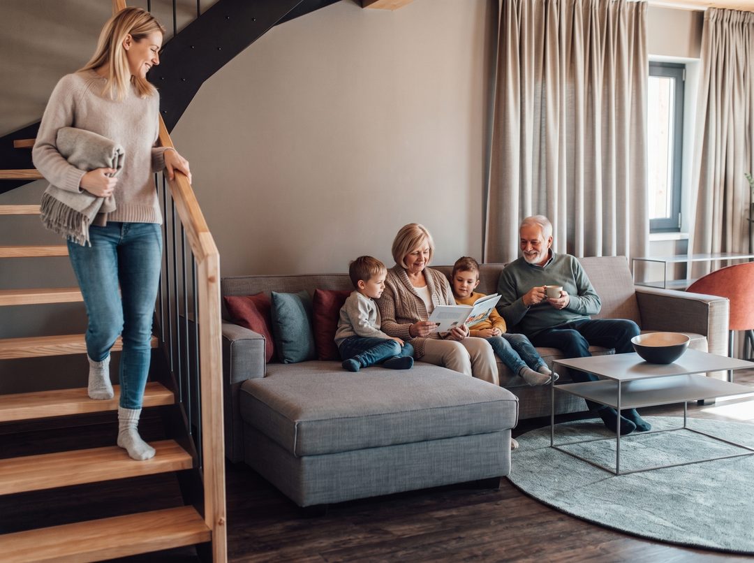 Drei Generationen im Wohnraum eines Chalets: Die Großmutter liest zwei Kindern vor, der Großvater trinkt Kaffee, die Mutter kommt mit einer Decke die Treppe herunter, Symbolbild