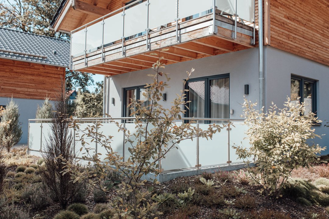Fachada de madera de un chalet con balcón y barandilla de cristal, con plantas en la terraza delante.