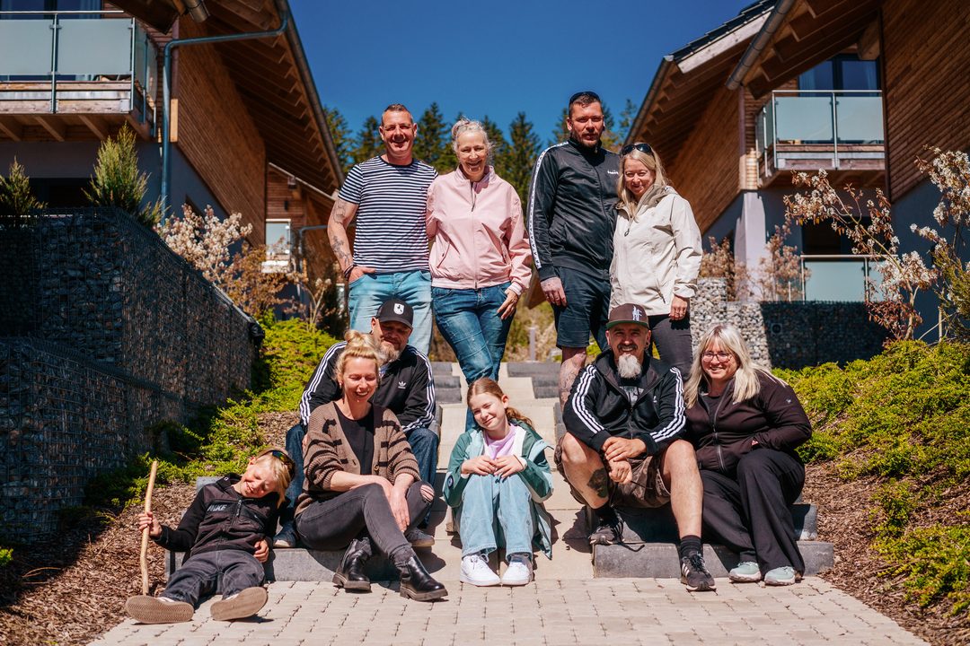 Ti gæster fra forskellige generationer sidder og står på den udendørs trappe mellem to træchalets i solen