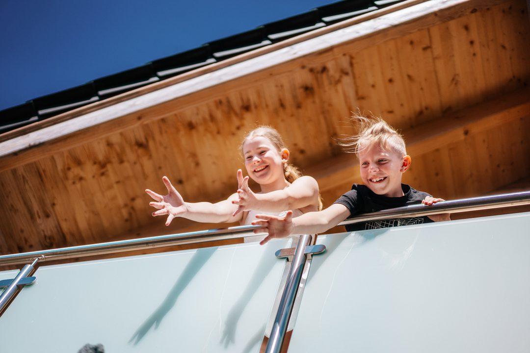 Dos niños sonrientes se asoman por la barandilla del balcón de un chalet, bajo el frontón de madera